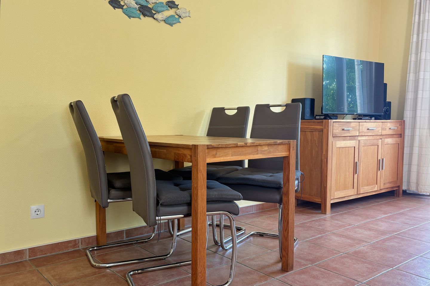 A dining area with a wooden table and chairs, a television on a cabinet, and a tiled floor.