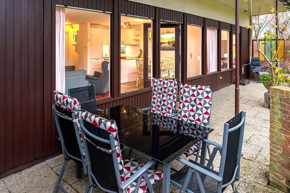 A dining area with a glass table and chairs, a kitchen visible through the open door, and a brick wall on one side. - (ULM1) Jürgens Lieblingsplatz Niendorf - Holiday home "Jürgens Lieblingsplatz (Timmendorfer Strand)