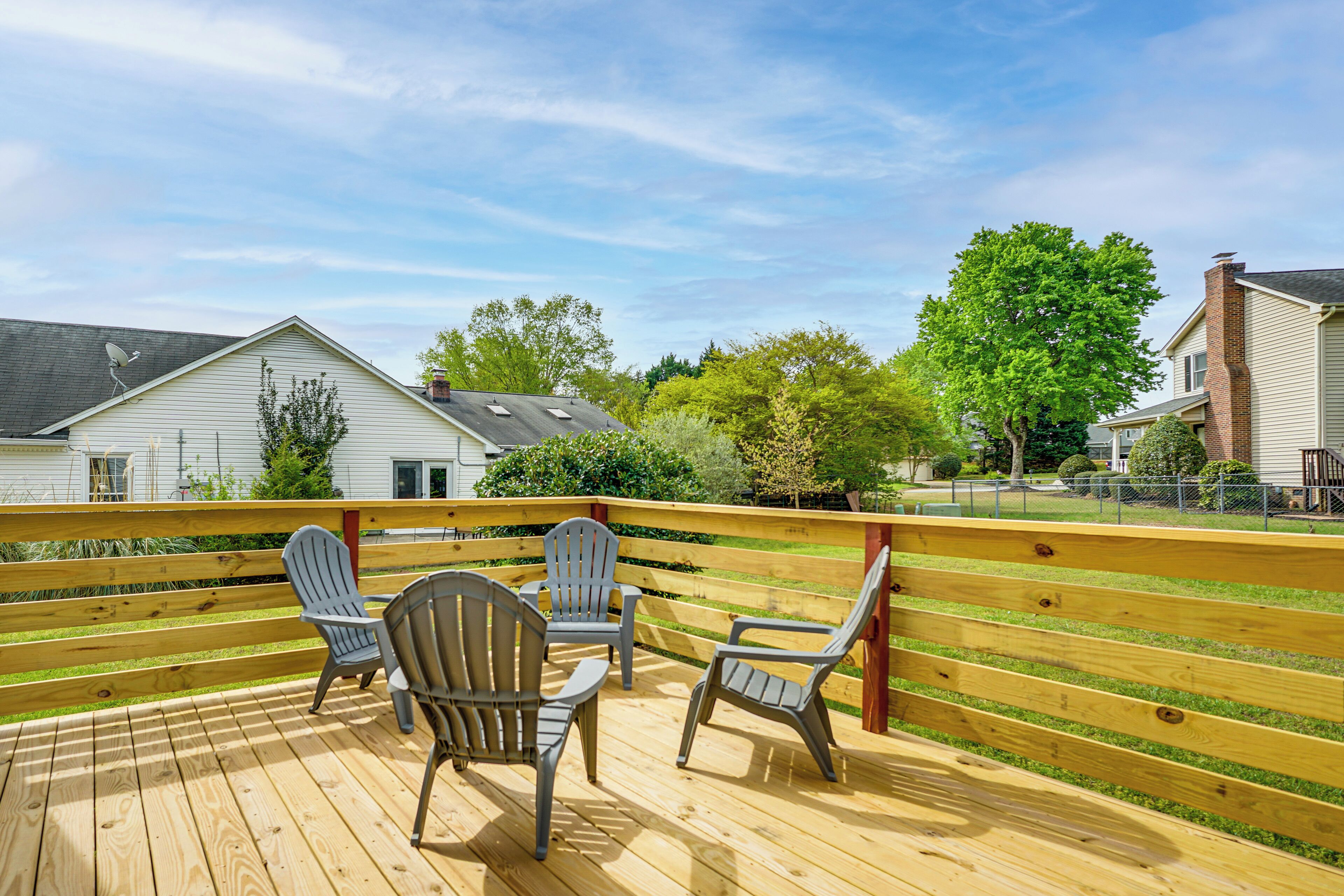 Een houten terras met vier Adirondack-stoelen, een leuning en uitzicht op huizen en bomen.