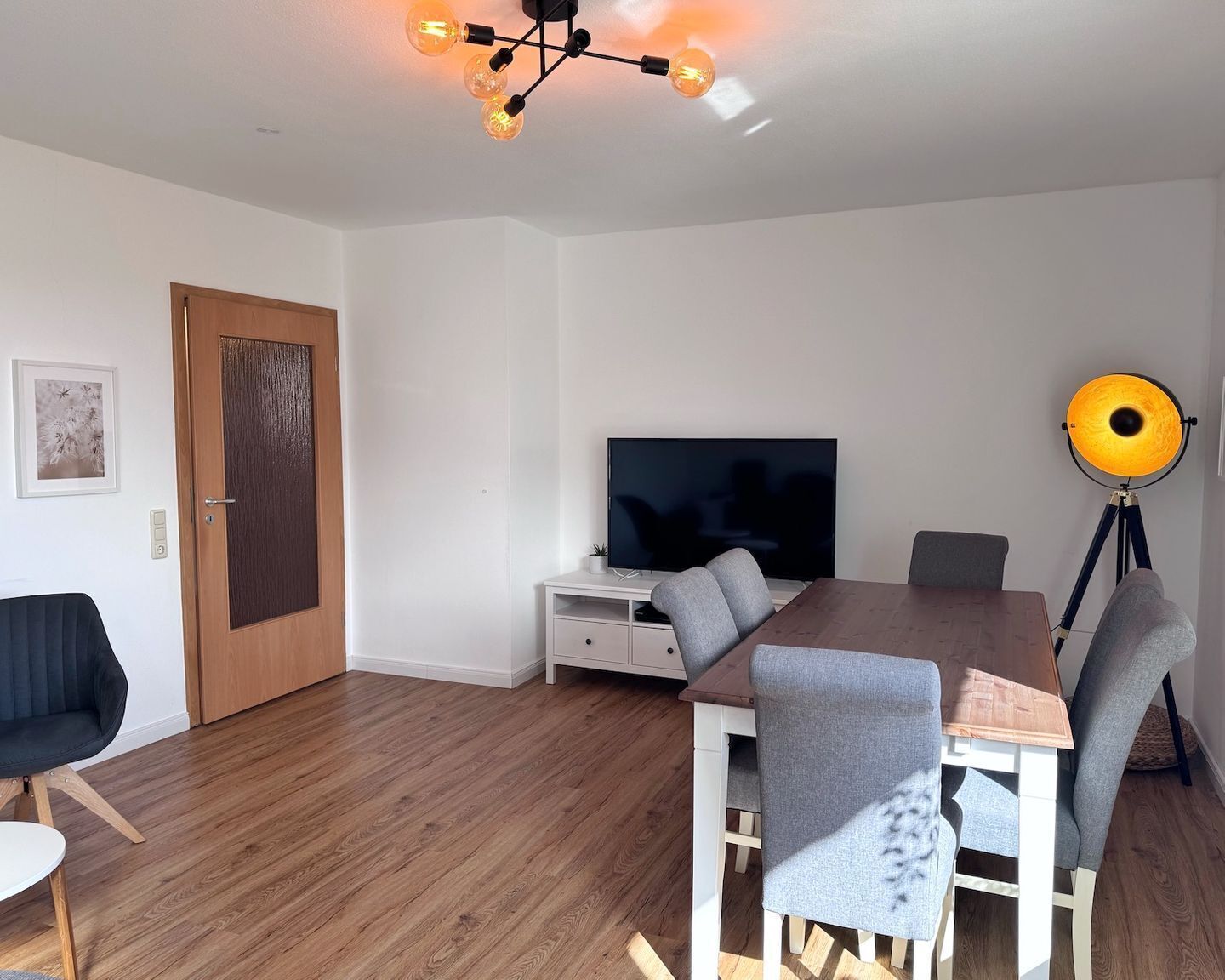 A modern living room with a wooden floor, a dining table with chairs, a flat-screen TV, a tripod lamp, and a door with a picture frame.