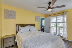 Un dormitorio con cama, mesita de noche, ventilador de techo y ventana con cortinas.