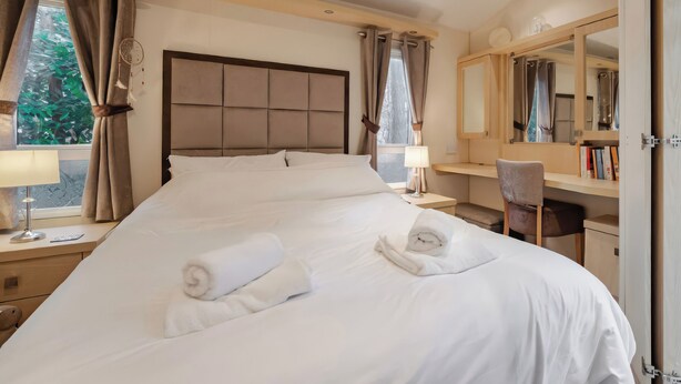 A hotel room with a large bed, a desk, a chair, a mirror, and a window with curtains. - Rawsons Retreat - -kelling Heath Holiday Park (Holt)
