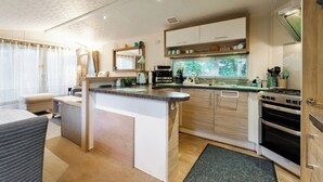 A modern kitchen with a central island, built-in oven, and a large window with a view. - Rawsons Retreat - -kelling Heath Holiday Park (Holt)