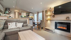 A modern living room with a sofa, a coffee table, a fireplace, a TV mounted on the wall, and a kitchen area in the background. - Rawsons Retreat - -kelling Heath Holiday Park (Holt)