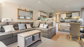 A modern living room with a sofa, a coffee table, and a dining area with a chair. - Rawsons Retreat - -kelling Heath Holiday Park (Holt)