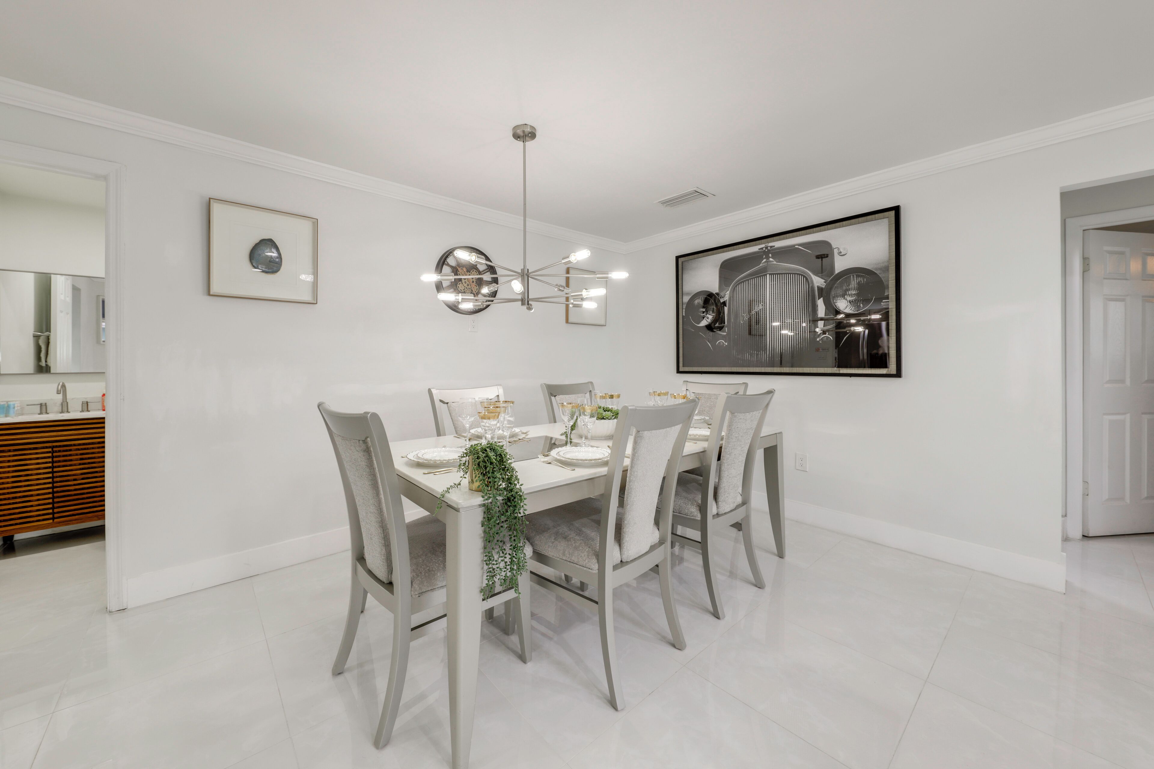 A dining area with a white table set for six, surrounded by chairs, a chandelier, and framed artwork on the walls.
