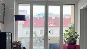 A room with a blue sofa, a small table, a floor lamp, and a sliding glass door leading to an outdoor area with a red-roofed building. - 120sqm Downtown luxury flat big S-side balcony (Wien)