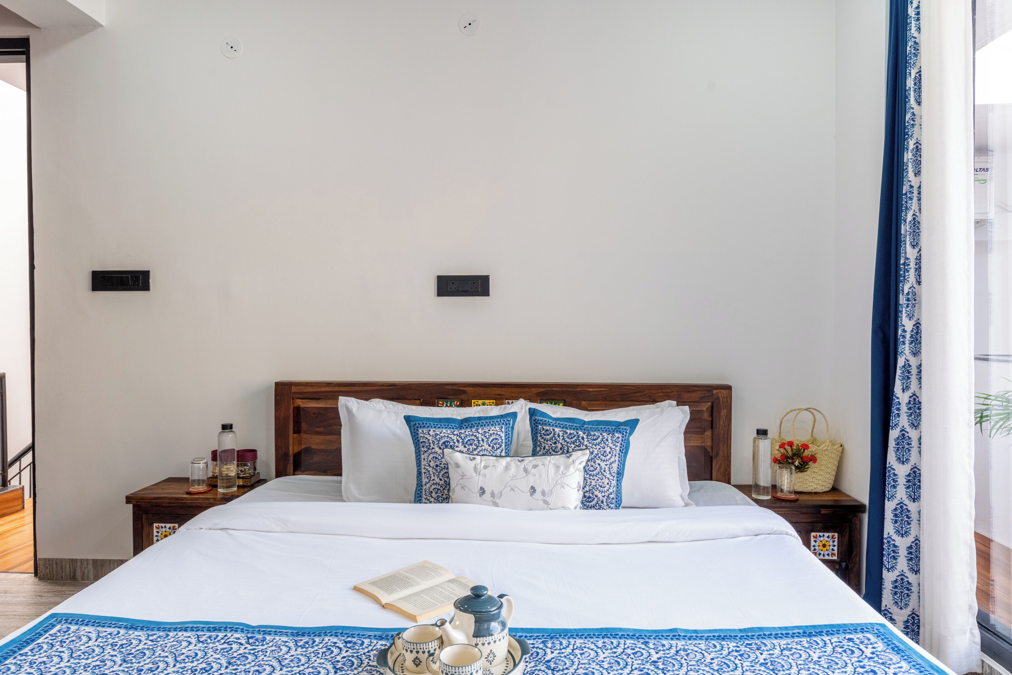 A neatly made bed with a blue and white patterned bedspread, a wooden headboard, and a bedside table with a lamp and a small basket.