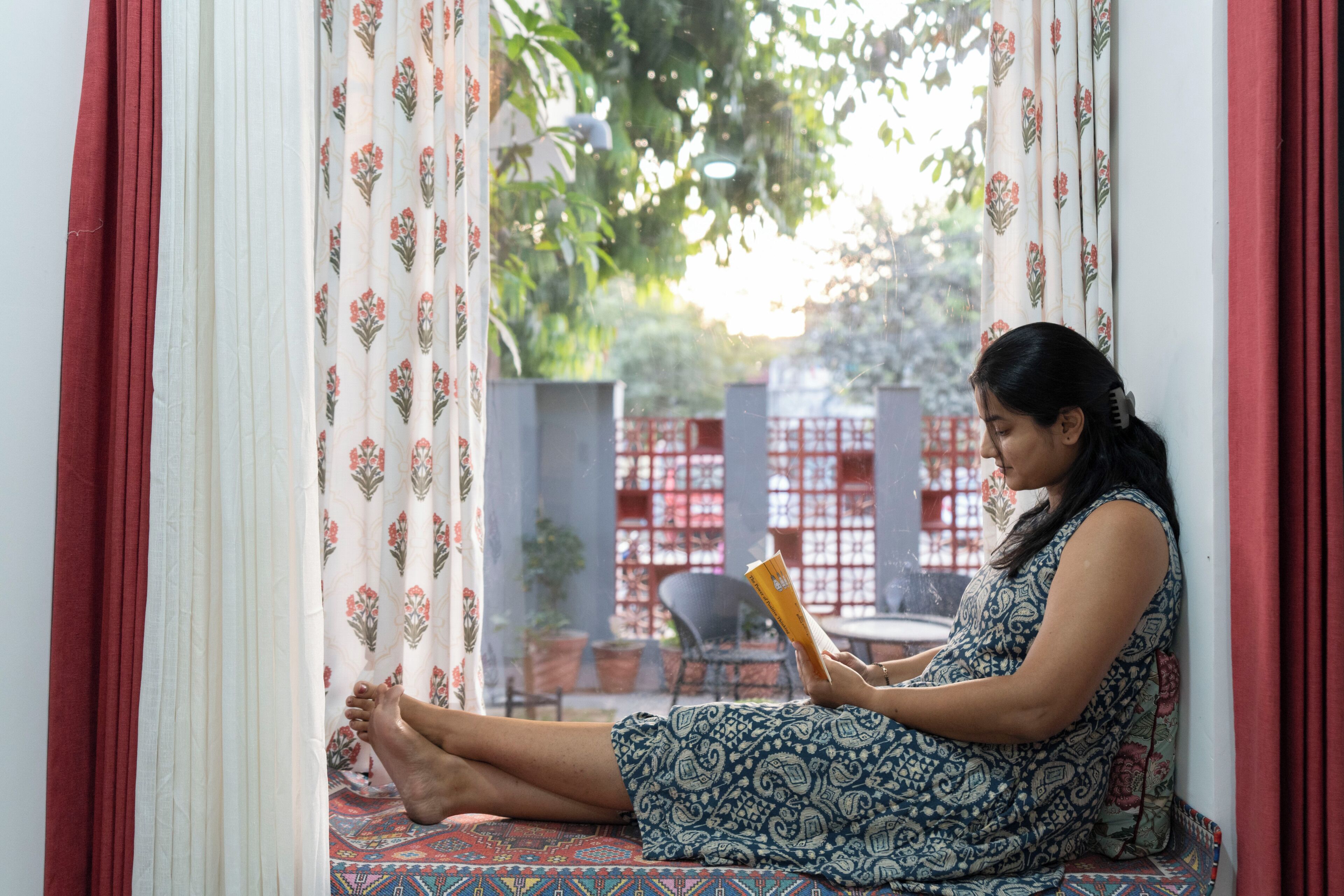 A person sitting on a patterned sofa by a window with curtains, reading a book.