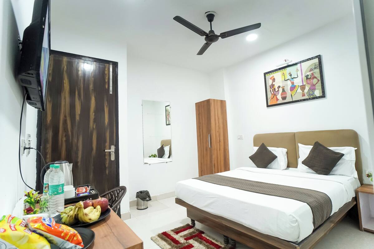 A hotel room with a bed, a ceiling fan, a wall-mounted TV, a wooden wardrobe, a mirror, and a small table with snacks.