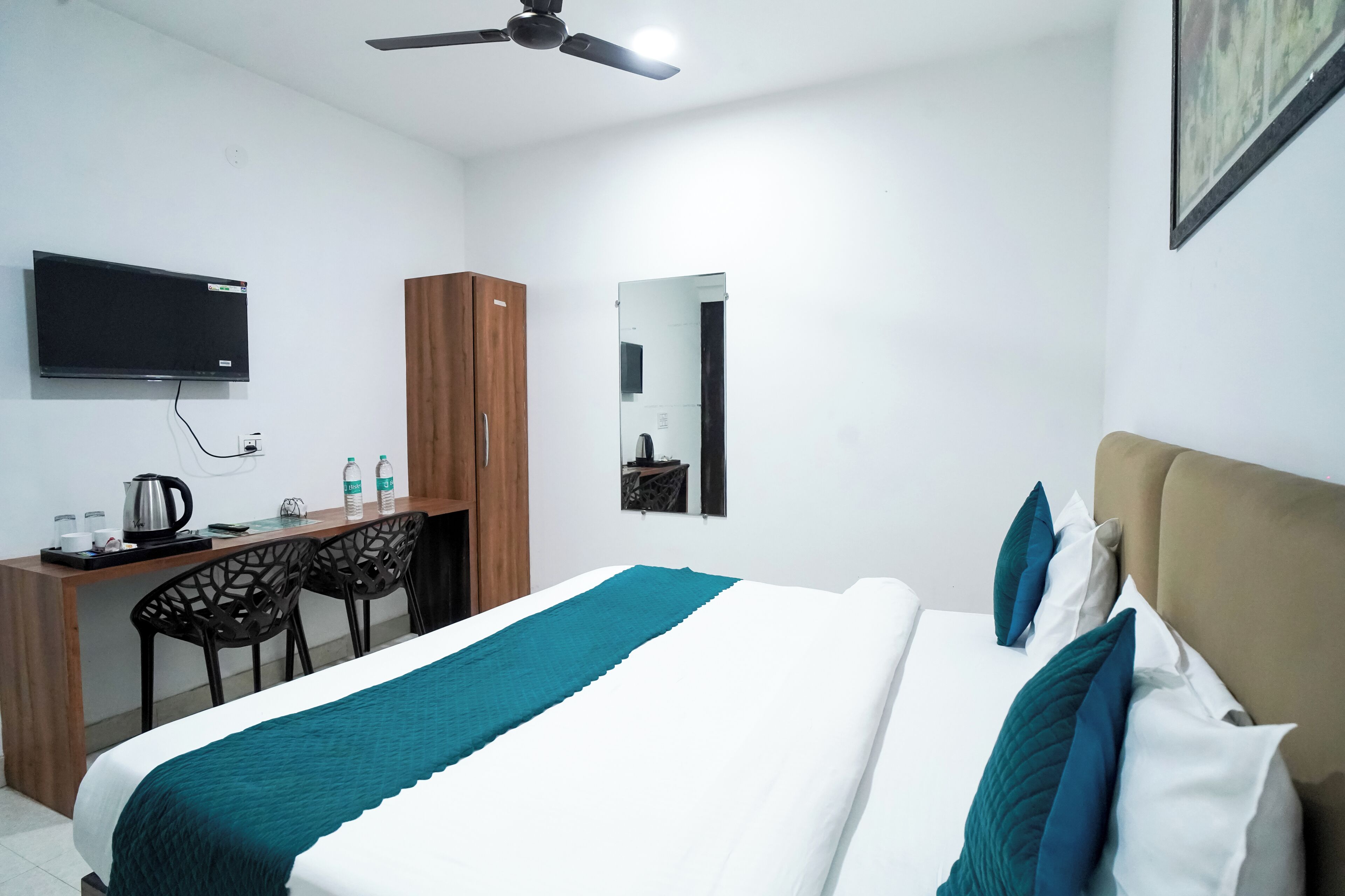 A hotel room with a bed, a desk with a television, a mirror, and a ceiling fan.