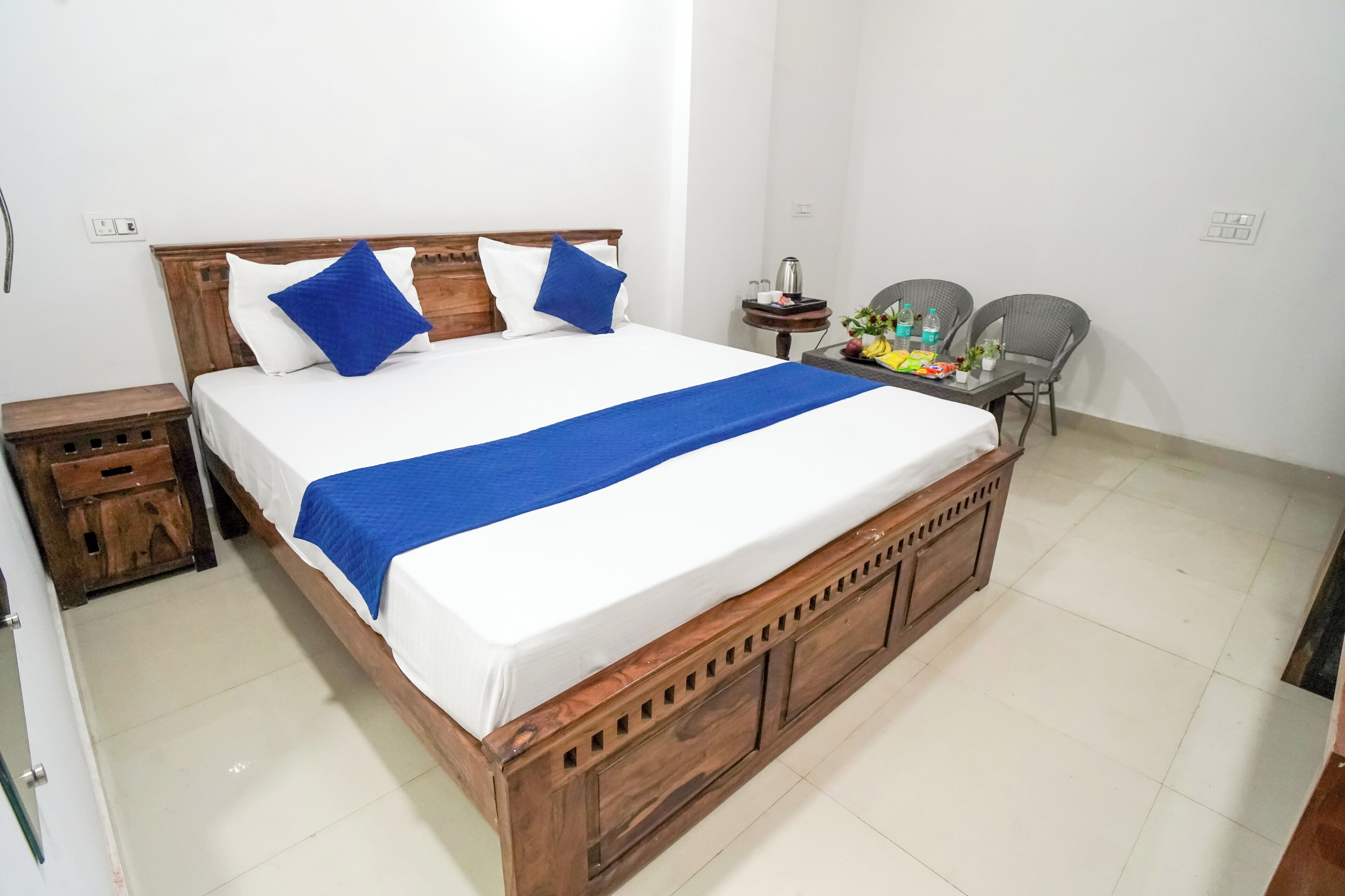 A bed with a wooden frame, a blue bedspread, and two blue pillows. There is a small table with a kettle and a fruit basket.
