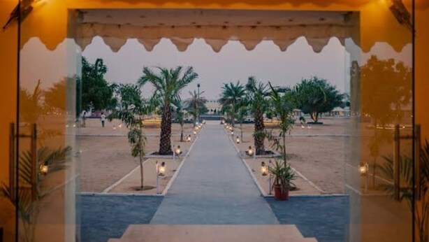 An outdoor pathway leading to a building entrance, flanked by palm trees and street lamps. - Desert Cactus Resort Jasialmer (Sam)