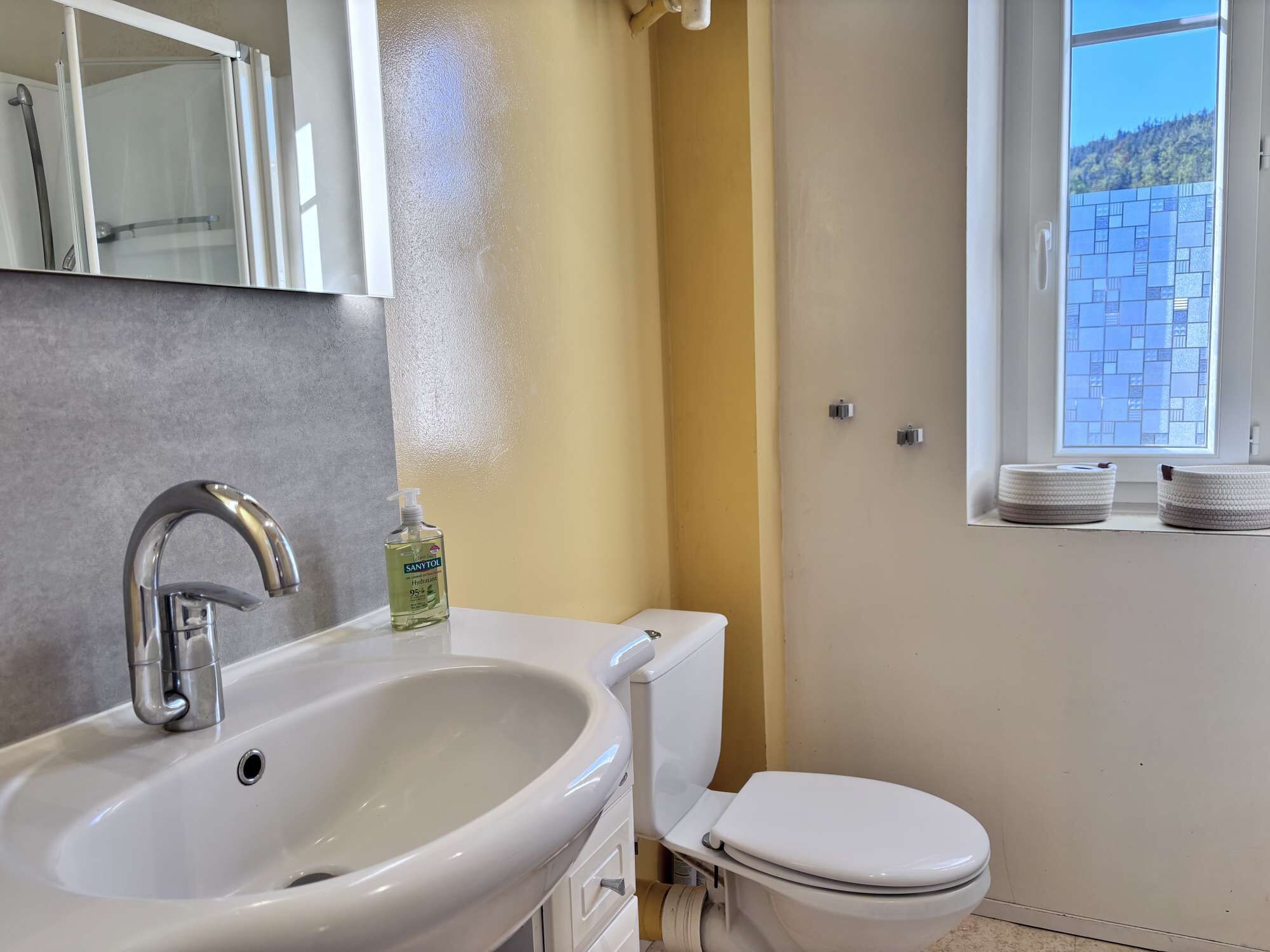 Une salle de bain avec un lavabo blanc, des toilettes et une fenêtre donnant sur un immeuble dont la façade est recouverte de carreaux bleus.