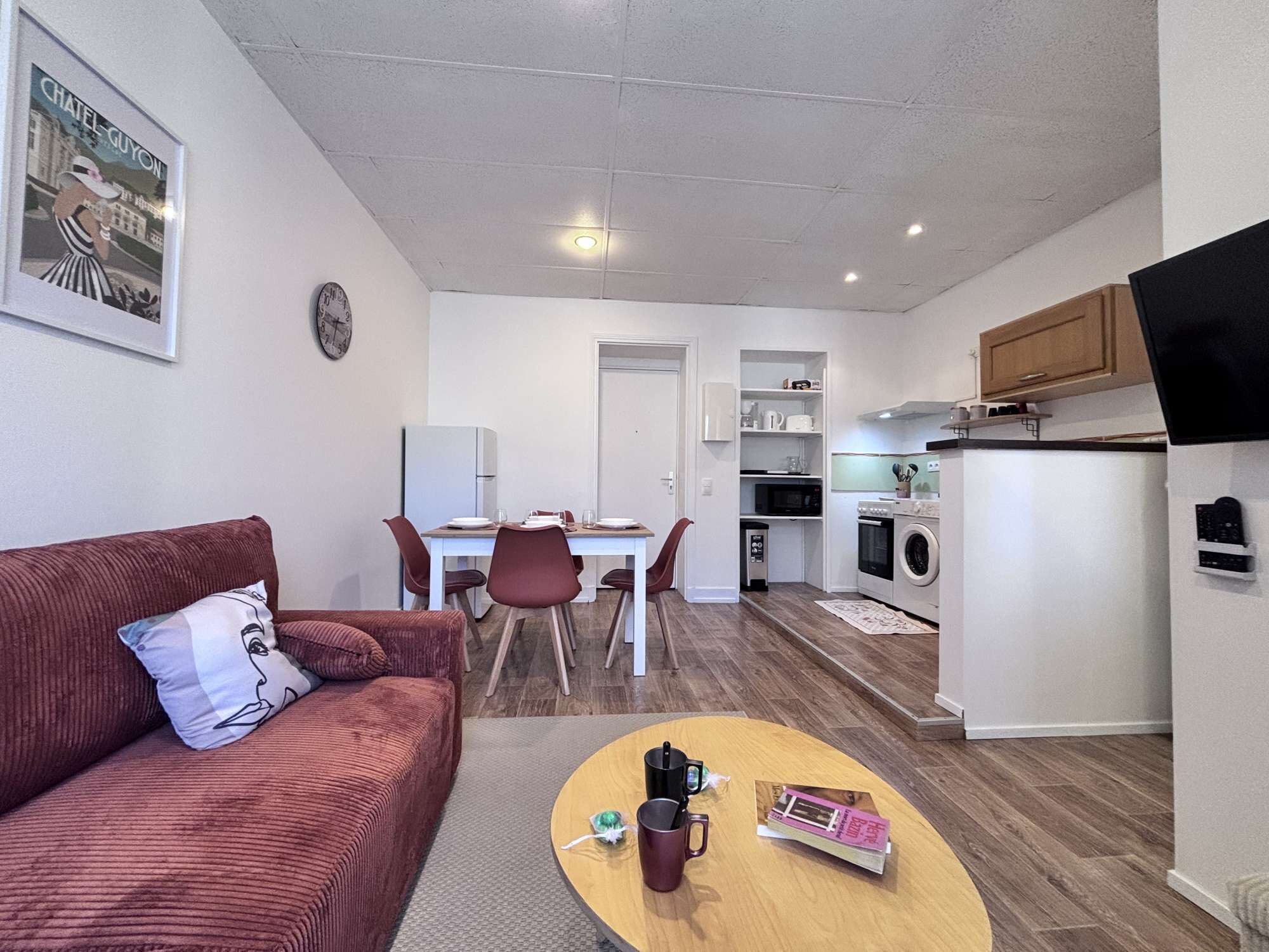 A compact living space with a sofa, a dining table, a kitchen area with appliances, and a TV mounted on the wall.