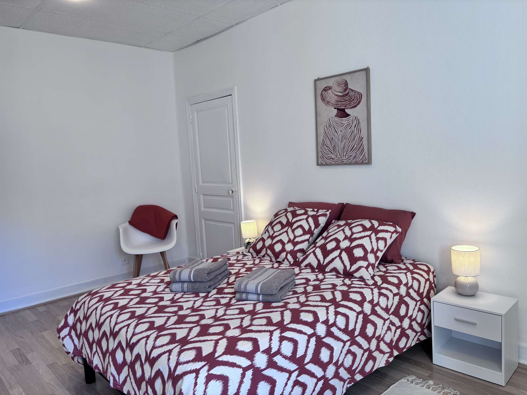 A bedroom with a patterned bedspread, a chair, a bedside table with a lamp, and a door.