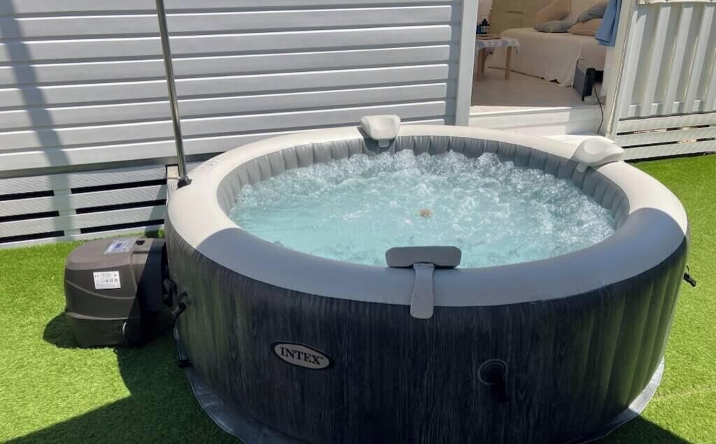 Un jacuzzi con bomba, rodeado de césped artificial, frente a una valla blanca y un edificio con revestimiento horizontal.
