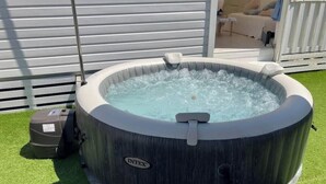 Ein Whirlpool mit Pumpe, umgeben von künstlichem Gras, vor einem weißen Zaun und einem Gebäude mit horizontaler Fassade.