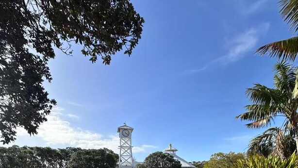 A tranquil water body with a clock tower, surrounded by lush greenery and a clear sky.