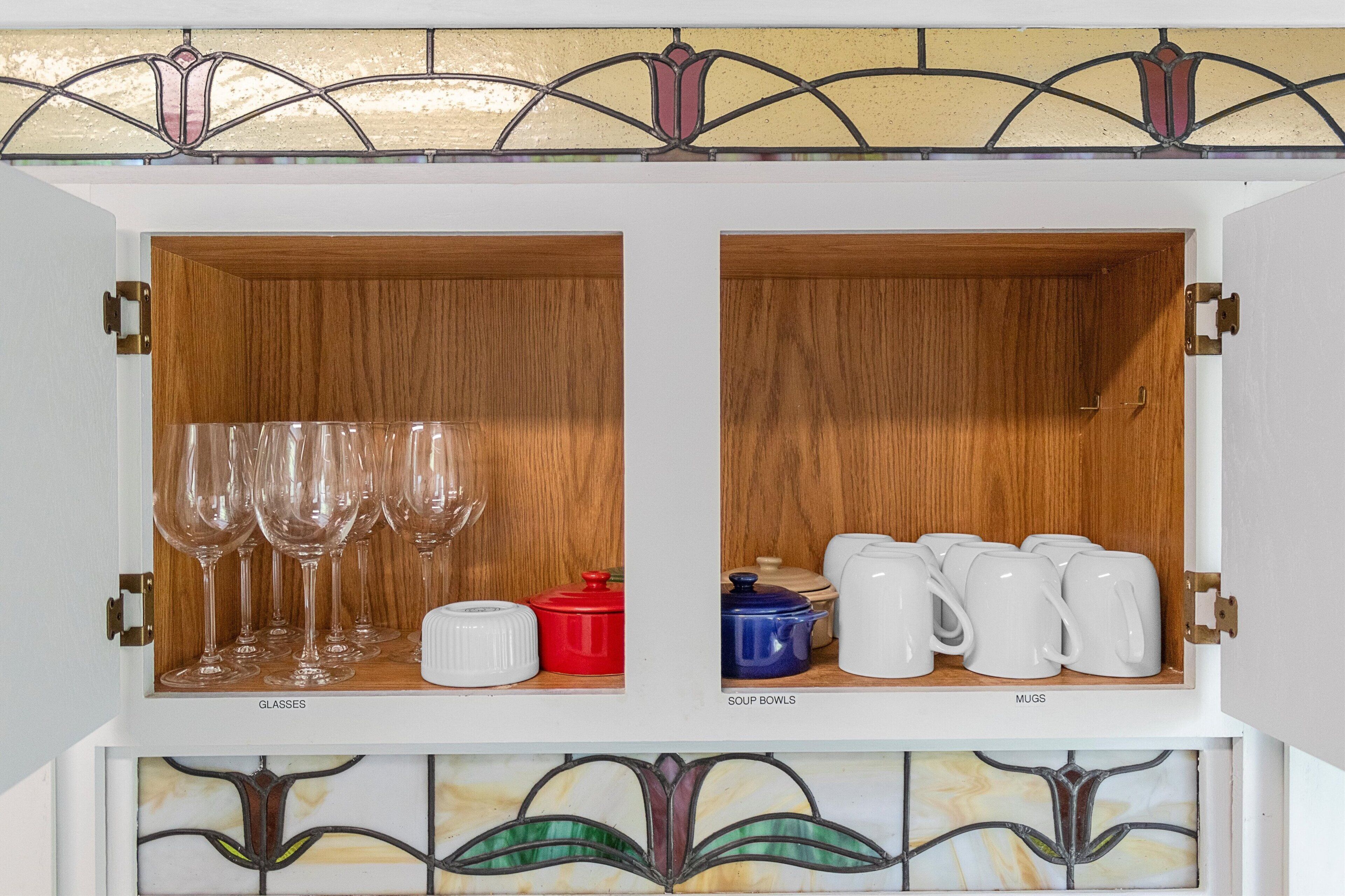 Ett skåp med två fack, det ena märkt med texten ”GLAS”, där det förvaras vinglas och en röd kastrull, och det andra märkt med texten ”SOPSKÅLAR”, där det förvaras vita muggar och en blå kastrull.