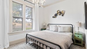 A bedroom with a large window, a chandelier, a bed with a headboard, two bedside tables, and wall decor.