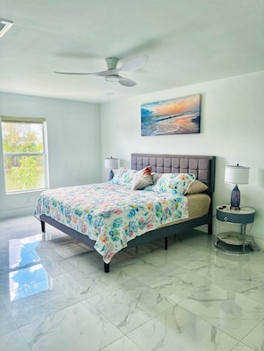 A bedroom with a bed, a ceiling fan, a painting, a window, and a nightstand. - Peaceful Waterfront Stay | Fishing Dock + Arcade (Cape Coral)