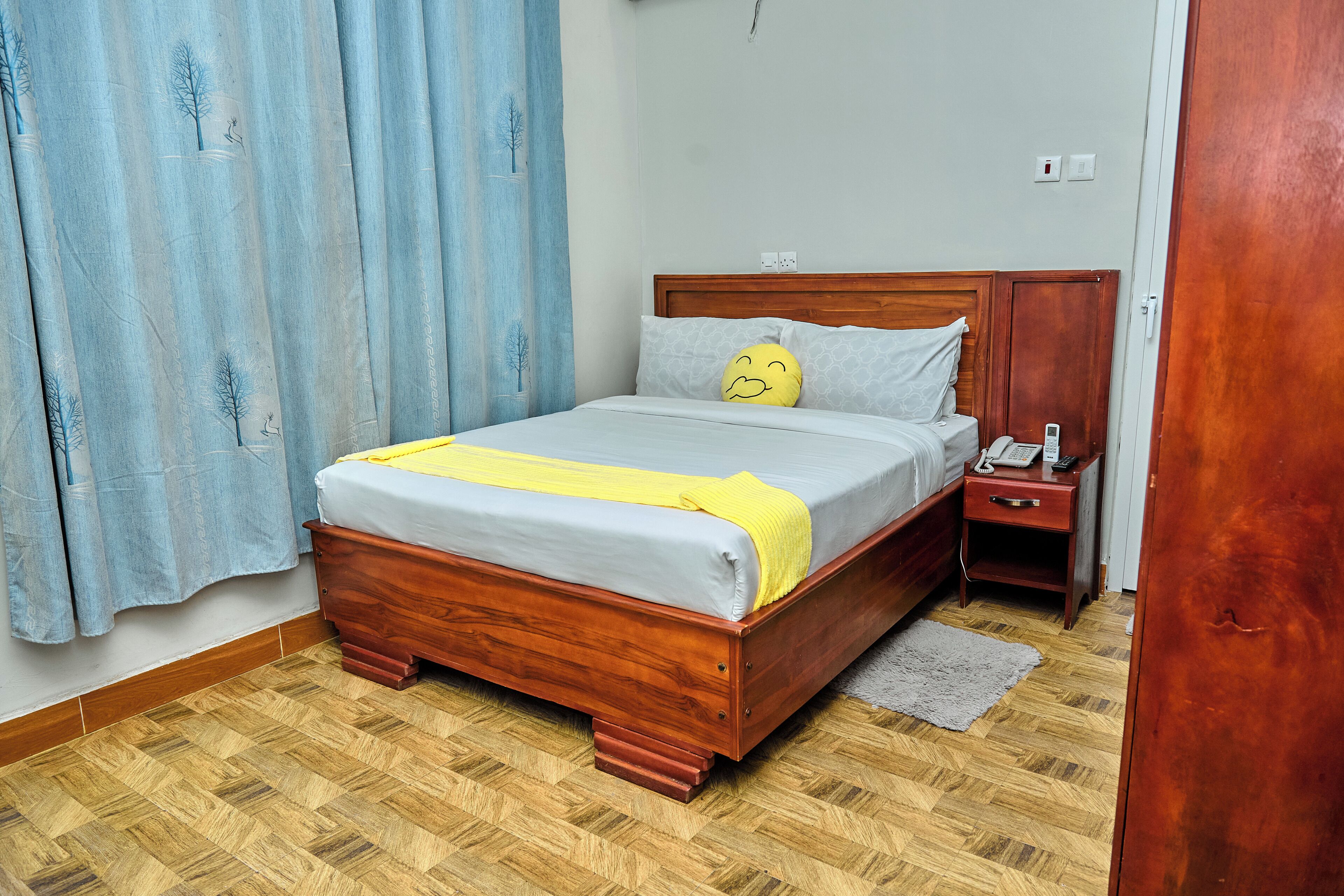 A bedroom with a wooden bed, a bedside table, a nightstand, a telephone, and a curtain.