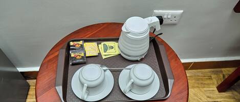 Una bandeja con dos tazas y platillos blancos, una tetera eléctrica blanca y algunos sobre de té sobre una mesa de madera.
