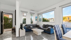 Ein modernes Wohnzimmer mit einer Couch, einem Couchtisch und einem Fernseher, großen Fenstern und einer Tür, die nach draußen führt.