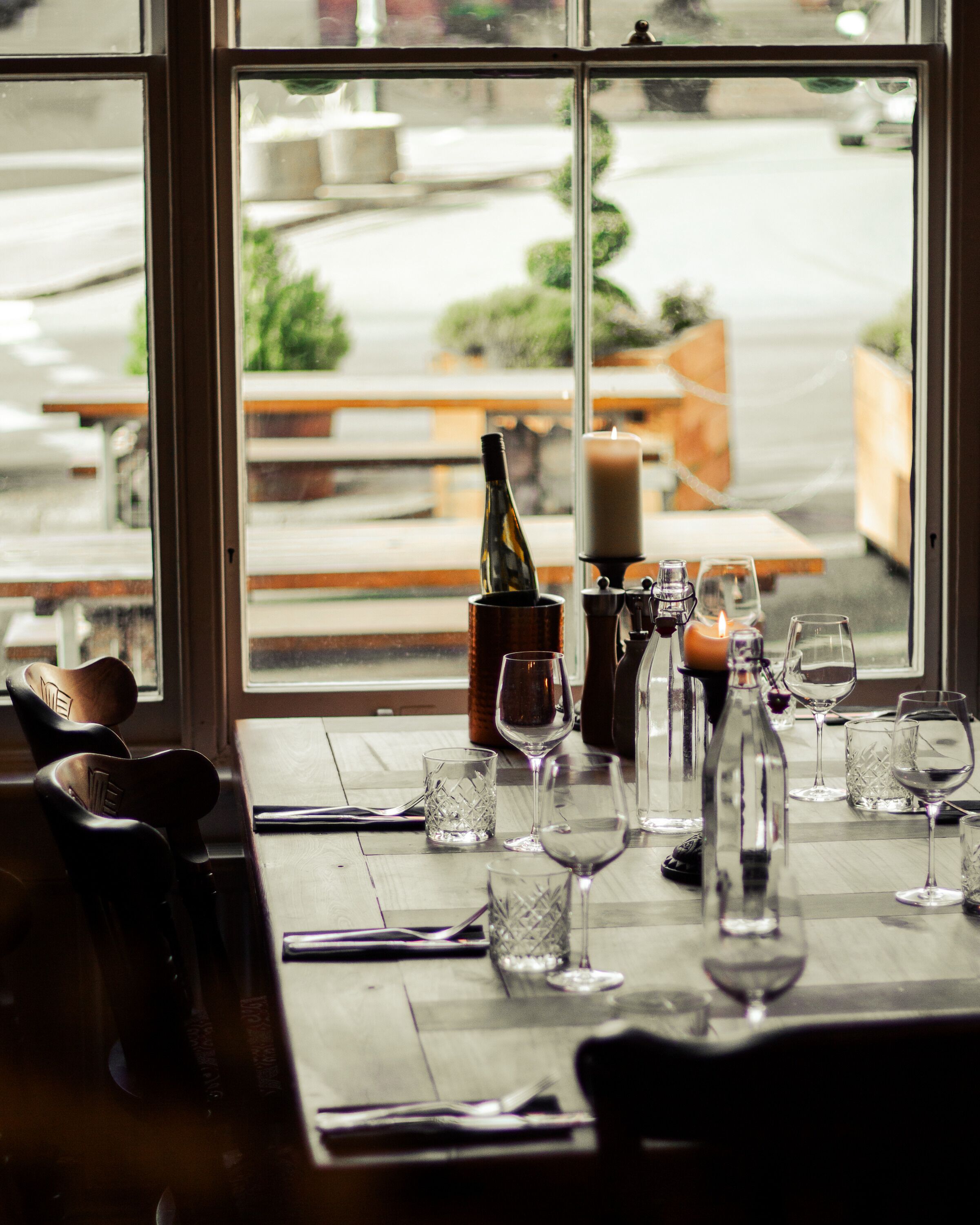 A restaurant table set with wine glasses, bottles, and a candle, with a view of the street outside.