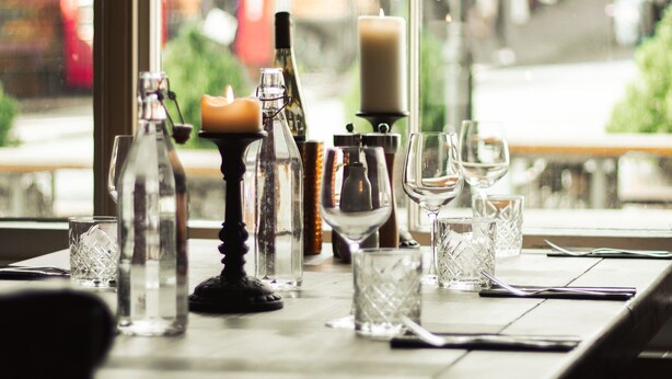 Une table dressée avec différents types de verres et de bouteilles, une bougie, et une vue par la fenêtre sur une scène de rue.