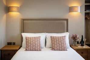 A neatly made bed with two pillows, a bedside table with a bottle and glasses, and a wall-mounted lamp. - The Bull Inn (Woodbridge)