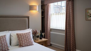 Une chambre d’hôtel comprenant un lit, une table de chevet, une lampe, une fenêtre avec des rideaux, un radiateur et un tableau encadré au mur.