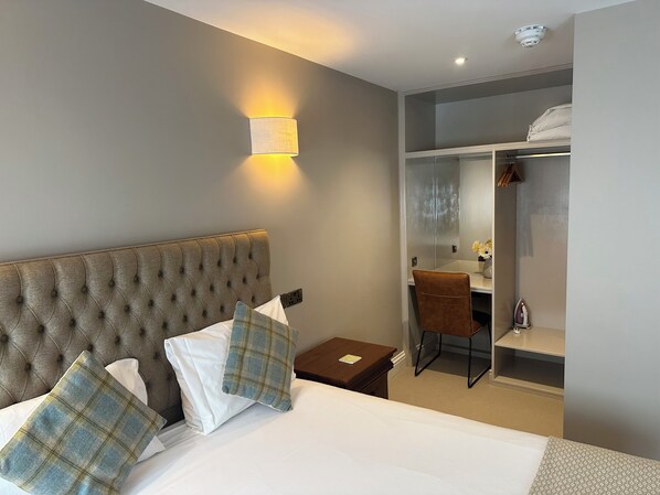 A hotel room with a tufted headboard, a bed with white linens, a plaid pillow, a small wooden nightstand, a desk with a chair, and a closet with a mirror. - The Bull Inn (Woodbridge)