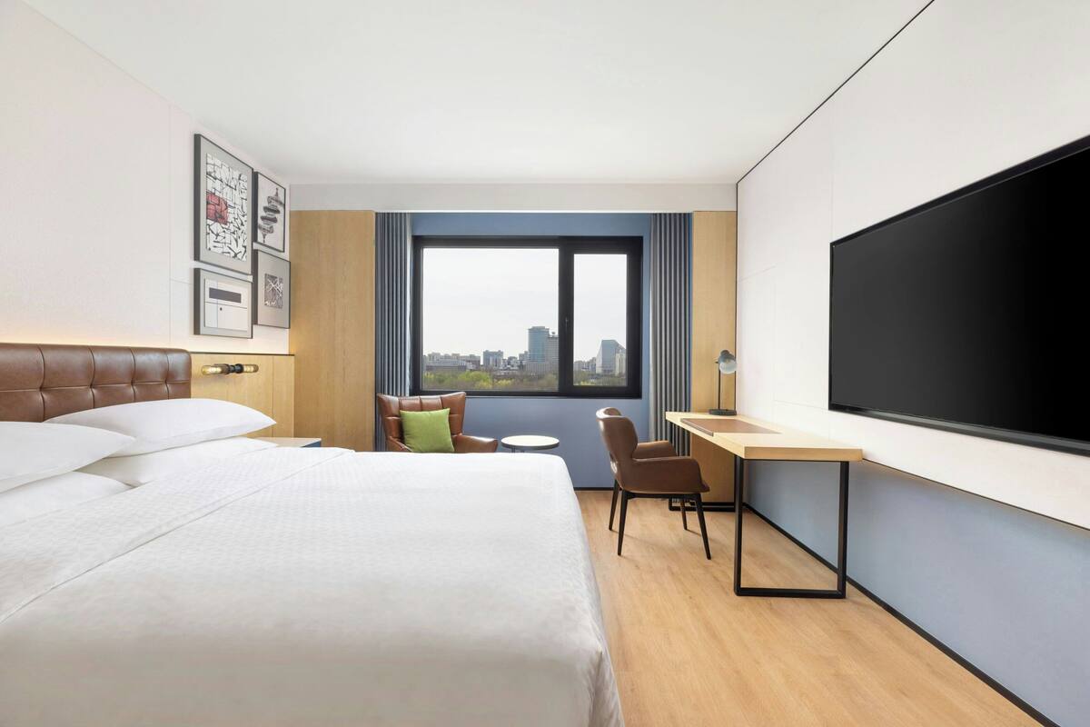 A hotel room with a bed, desk, chair, large window, and a flat-screen TV.