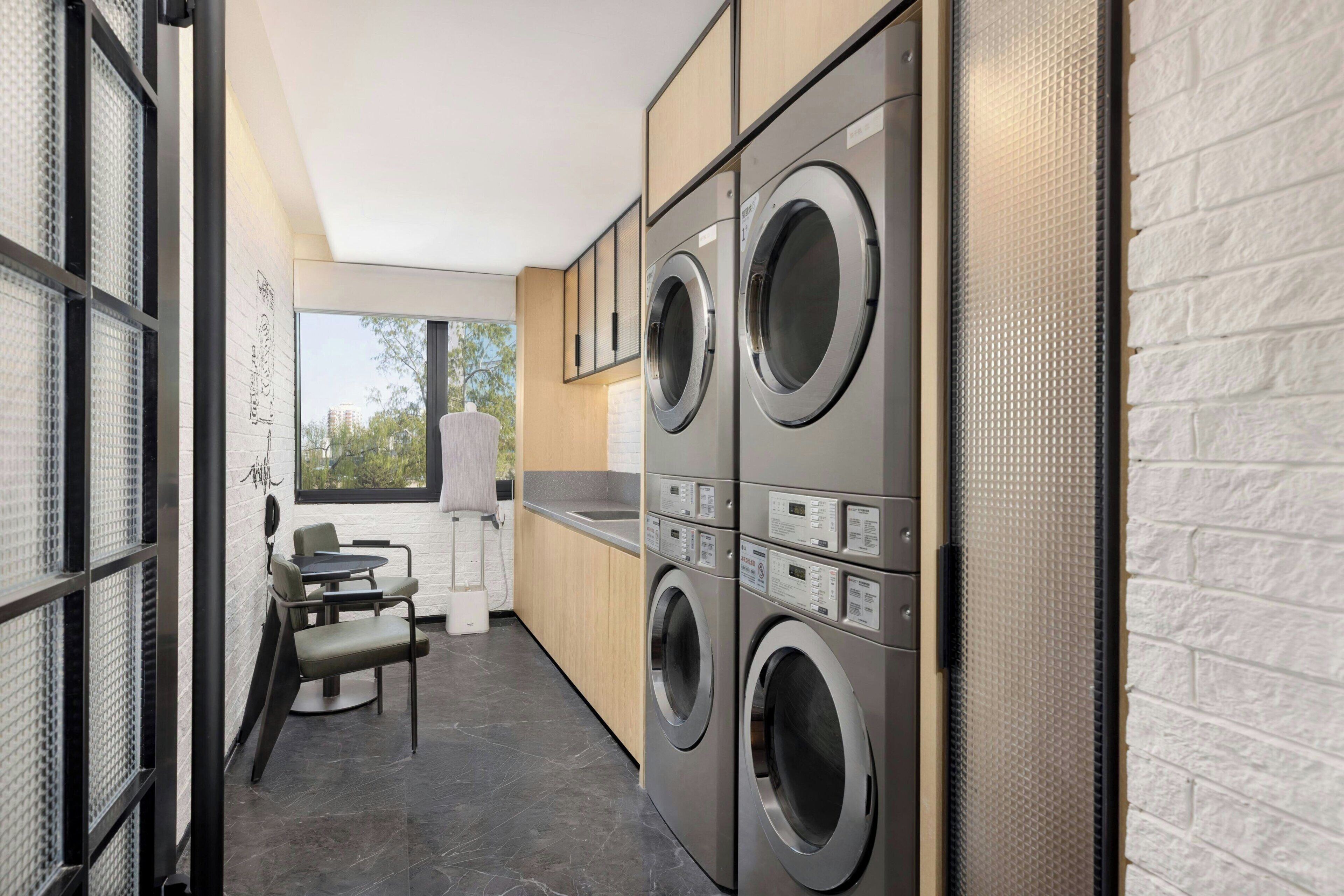 A modern laundry room with stacked washers and dryers, a small seating area with chairs, and a view through a window.