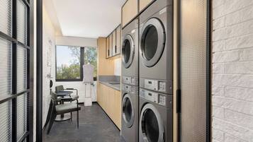 A modern laundry room with stacked washers and dryers, a small seating area with chairs, and a view through a window.