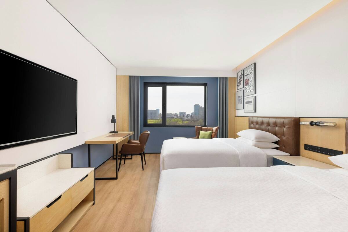 A hotel room with a large bed, a desk, a chair, a TV, and a window with a city view.