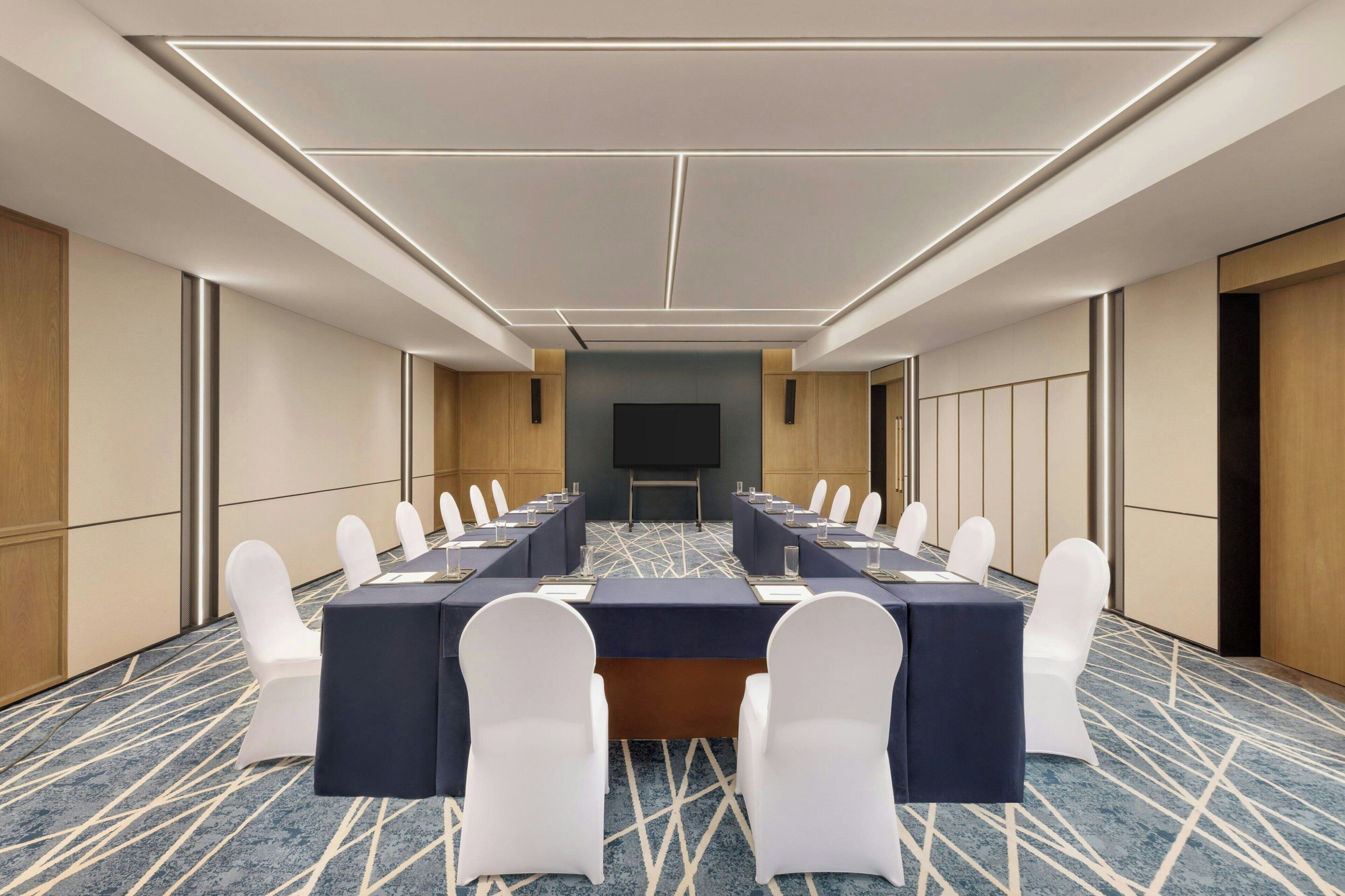 A conference room with a long table, white chairs, and a large screen.