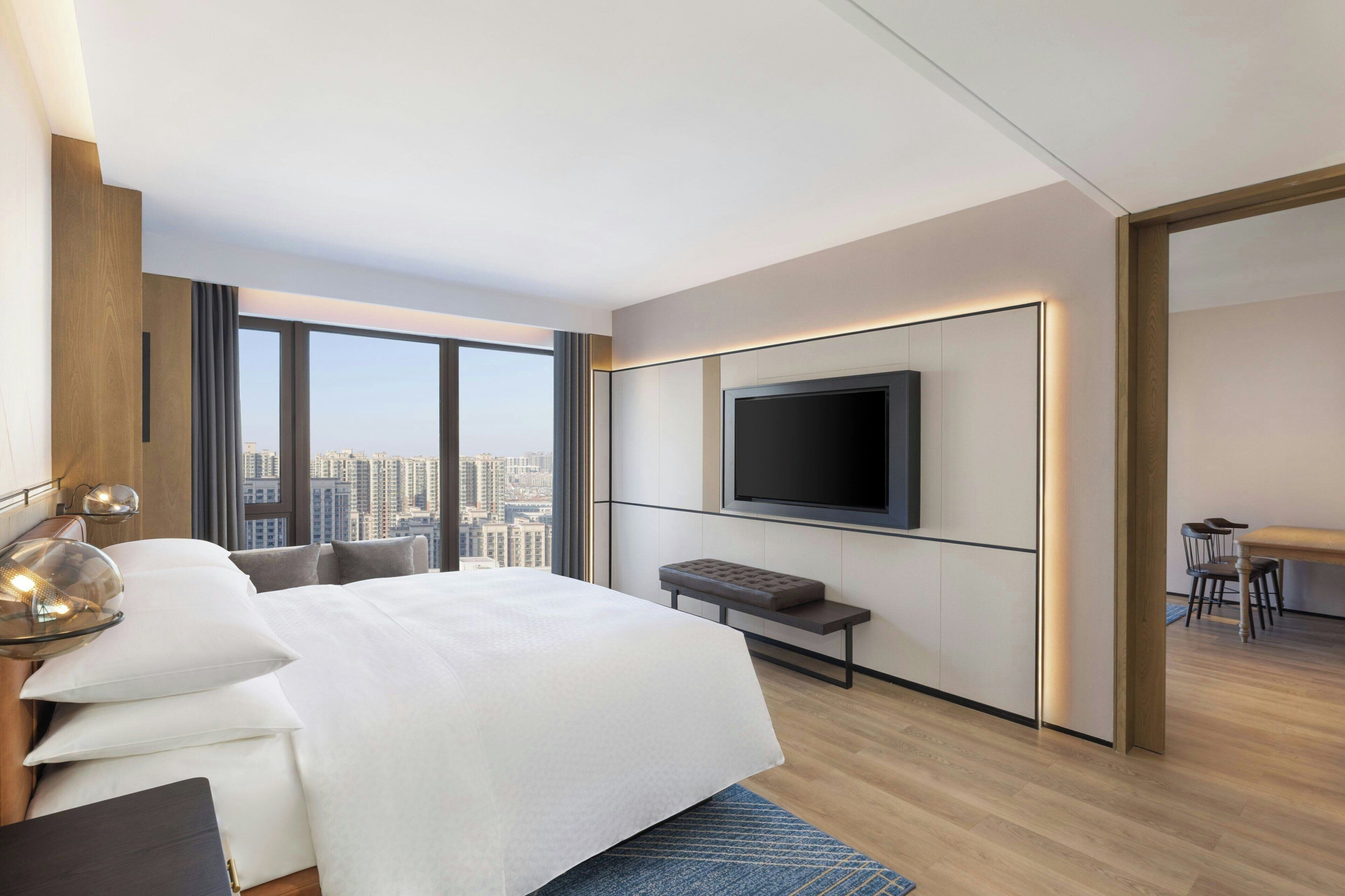A modern hotel room with a large bed, a TV mounted on the wall, a city view, and a dining area.