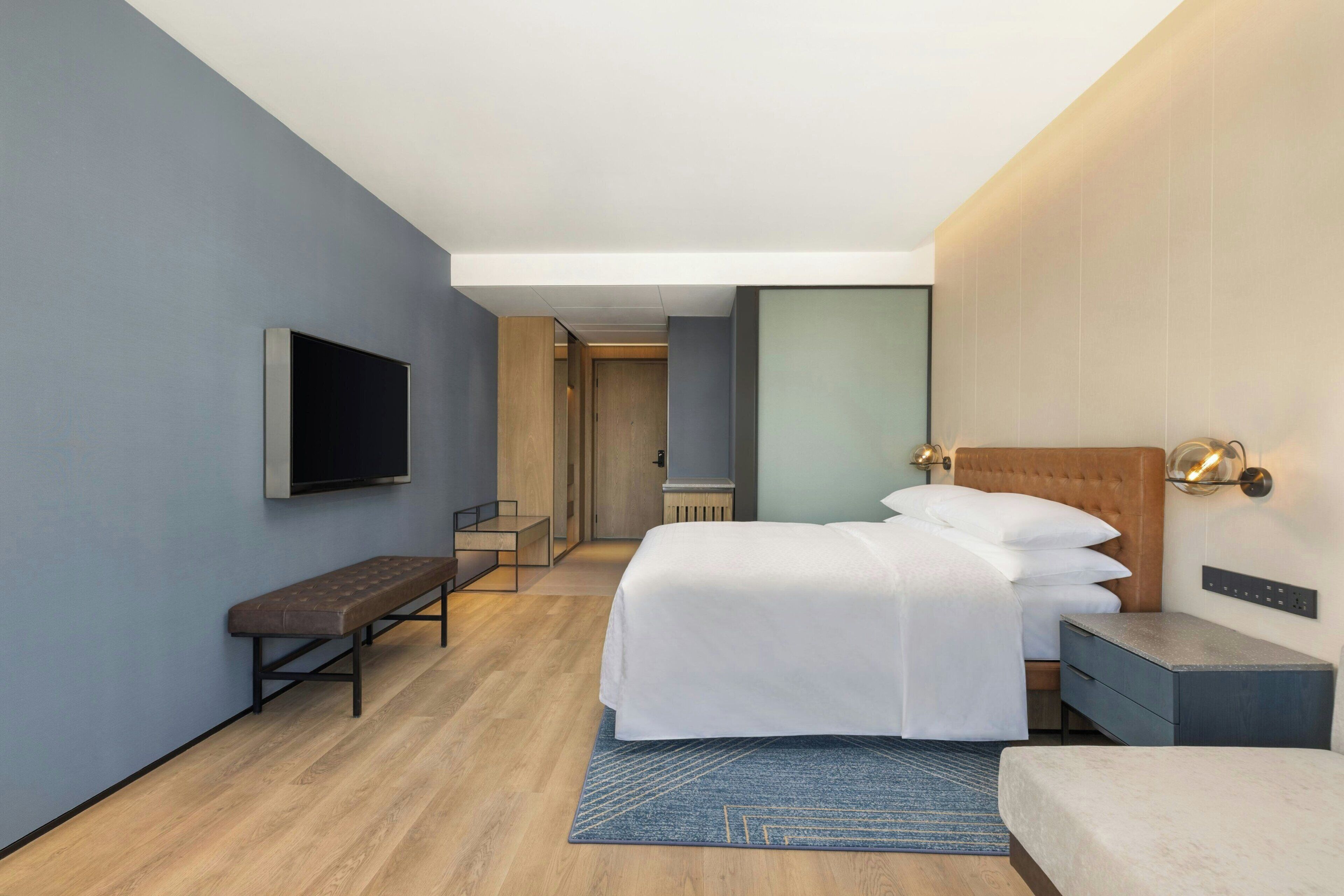 Una habitación de hotel moderna con cama, banco, mesita de noche y televisor de pantalla plana.