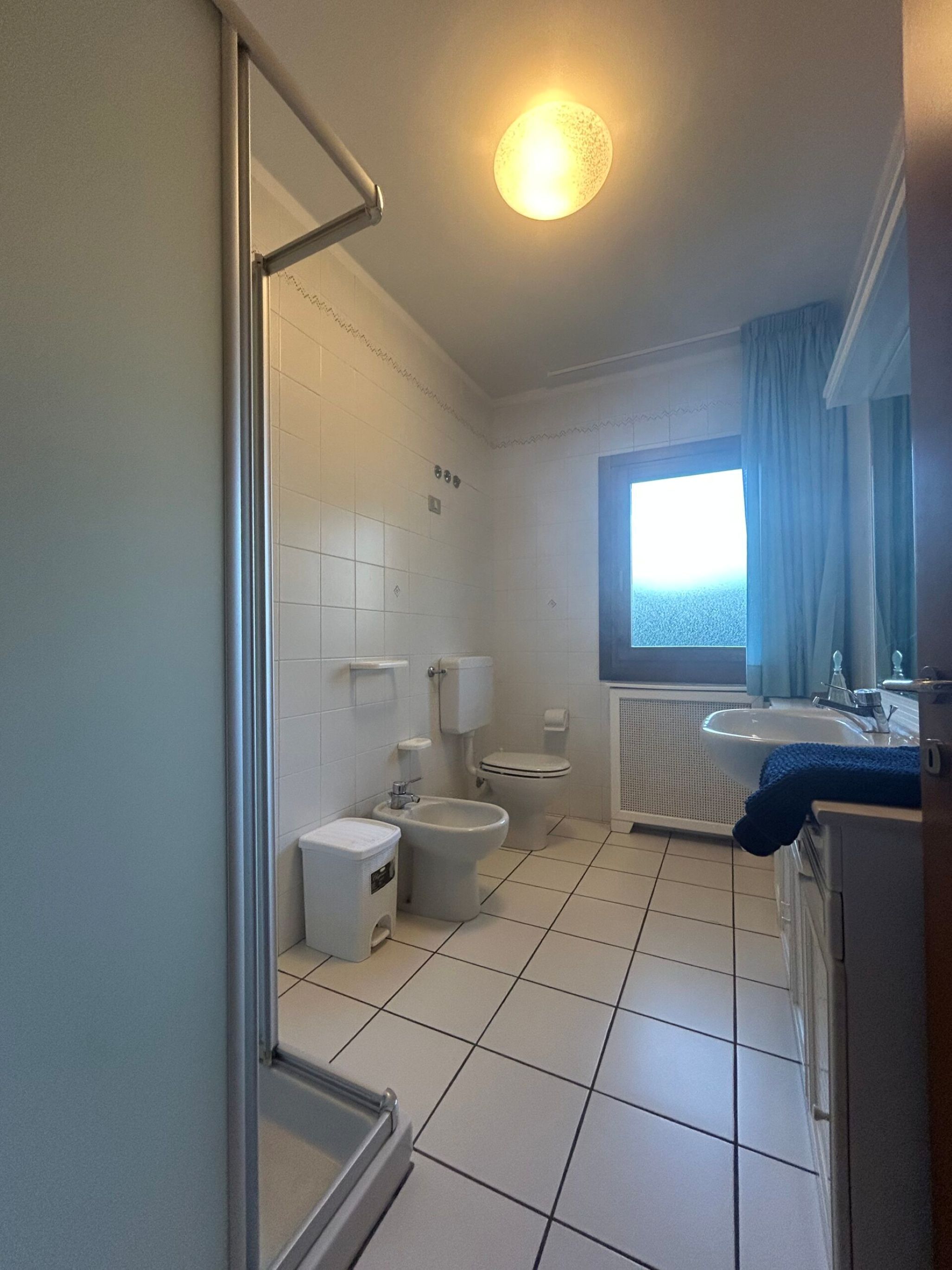 A bathroom with a toilet, bidet, and sink, a window with curtains, and a shower area.