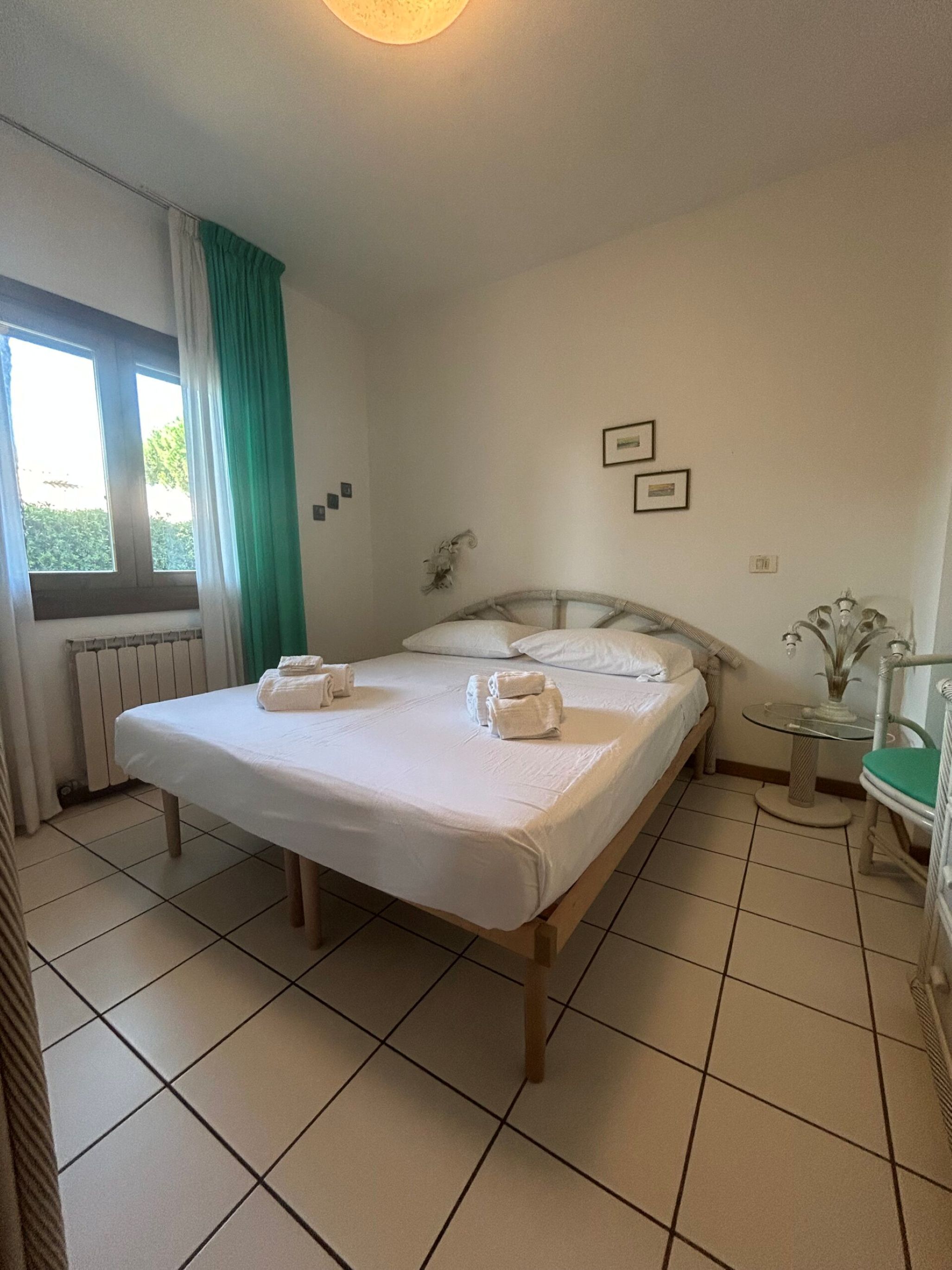 A neatly made bed with towels, a window with curtains, a radiator, and a small table with a plant.