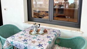 Une petite table recouverte d’une nappe à motifs d’étoiles de mer, deux chaises vertes et une fenêtre donnant sur des bâtiments et des arbres.