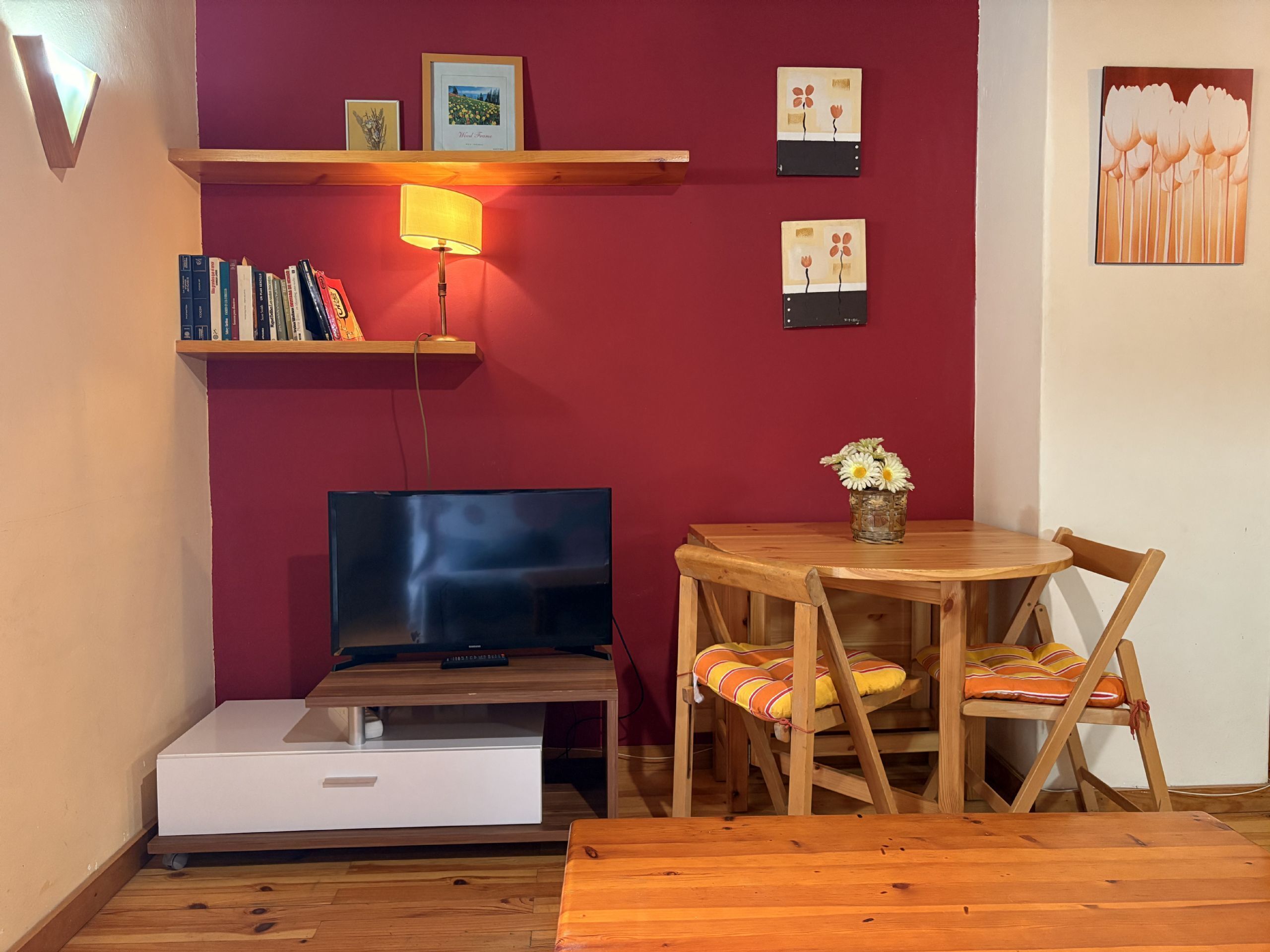 Una habitación con una televisión sobre un soporte de madera, una mesita con un florero y una pared roja con cuadros enmarcados.