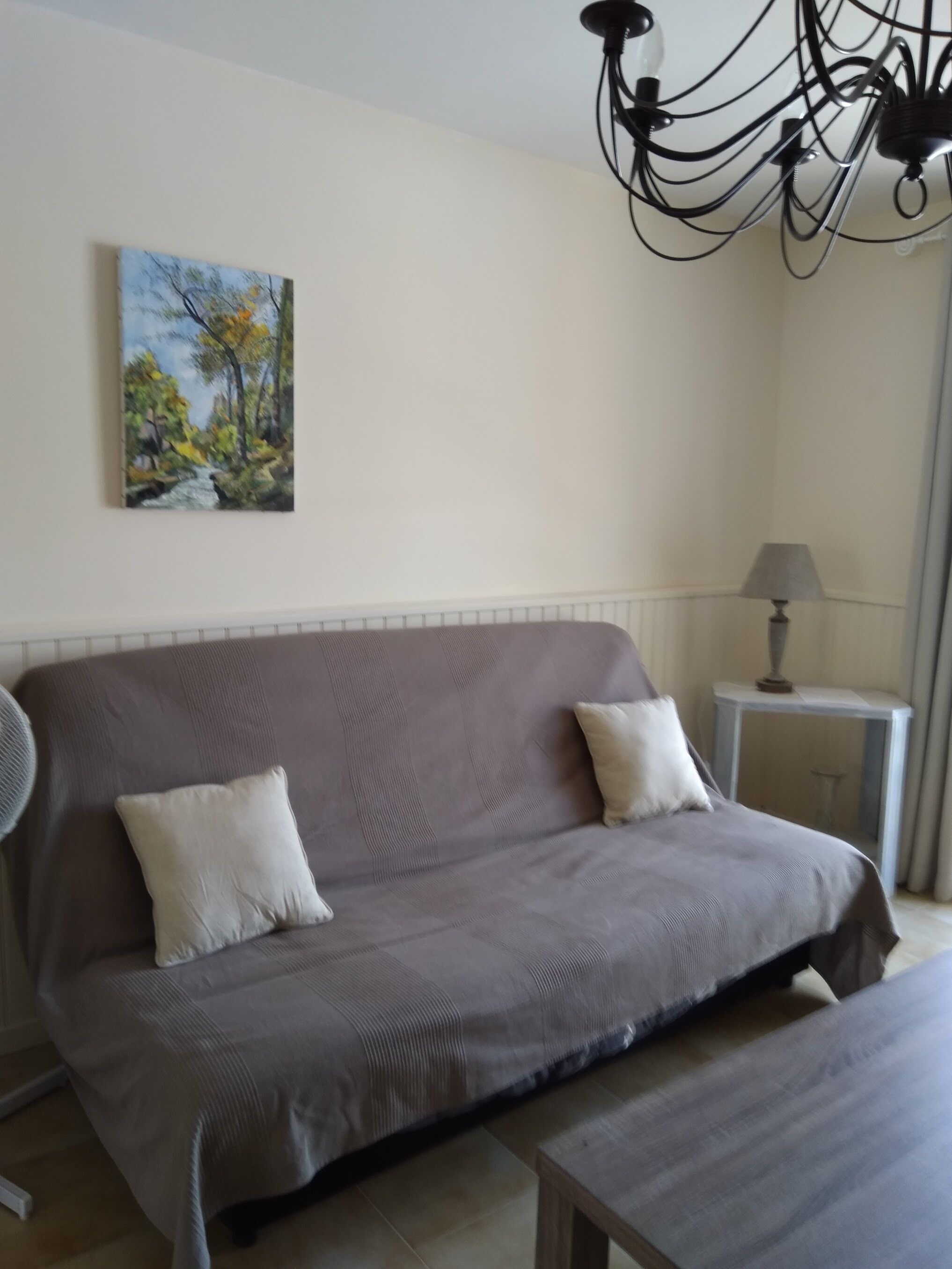 Una habitación con sofá, dos almohadas, lámpara, ventilador y un cuadro en la pared.