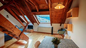 Een kamer met een schuine balkenplafond, een tafel met een plant, een stoel, een radiator en een raam met uitzicht.