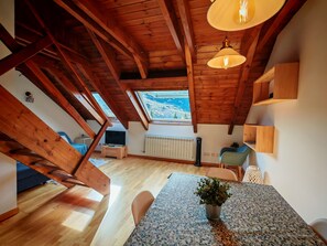 Una habitación con techo inclinado, vigas de madera, una mesa con una planta, una silla, un radiador y una ventana con vistas.