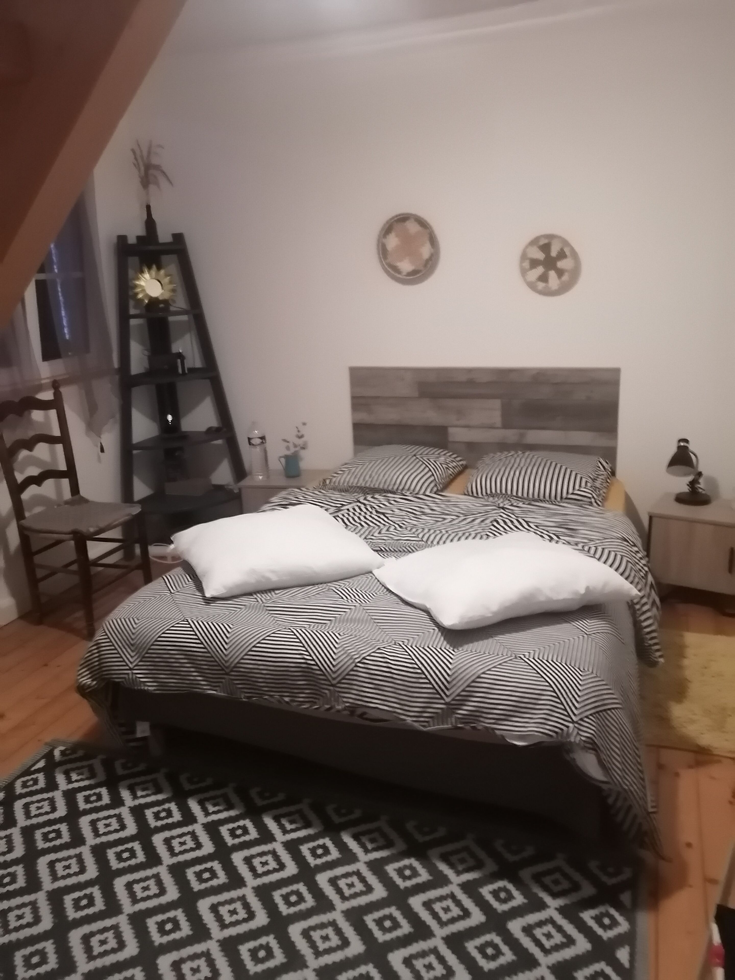 Une chambre à coucher comprenant un lit, une chaise en bois, une étagère avec des objets décoratifs et un tapis à motifs.