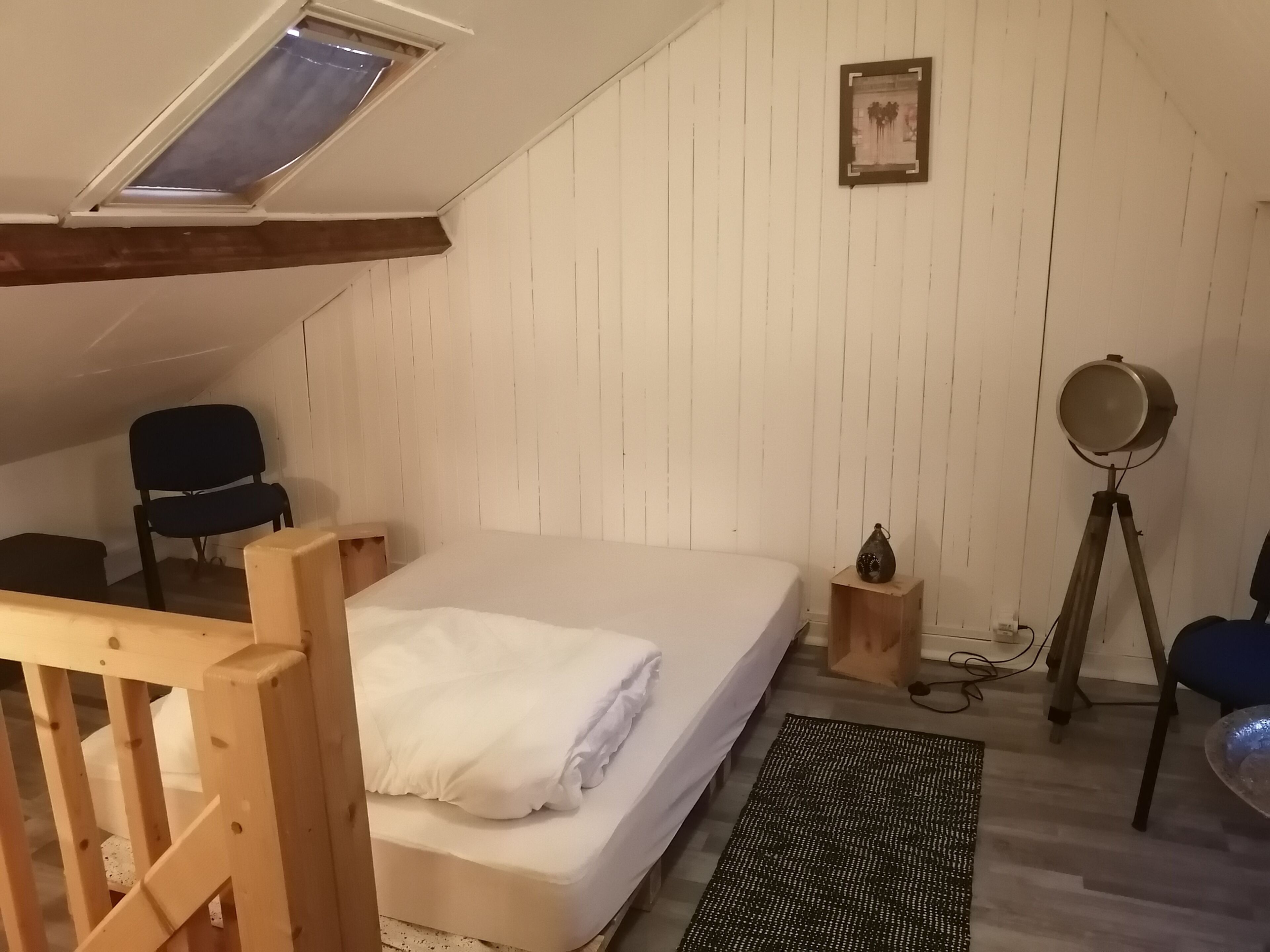Huone, jossa on kalteva katto, sänky, tuoli, pieni pöytä, lattiavalaisin ja taulu seinällä.